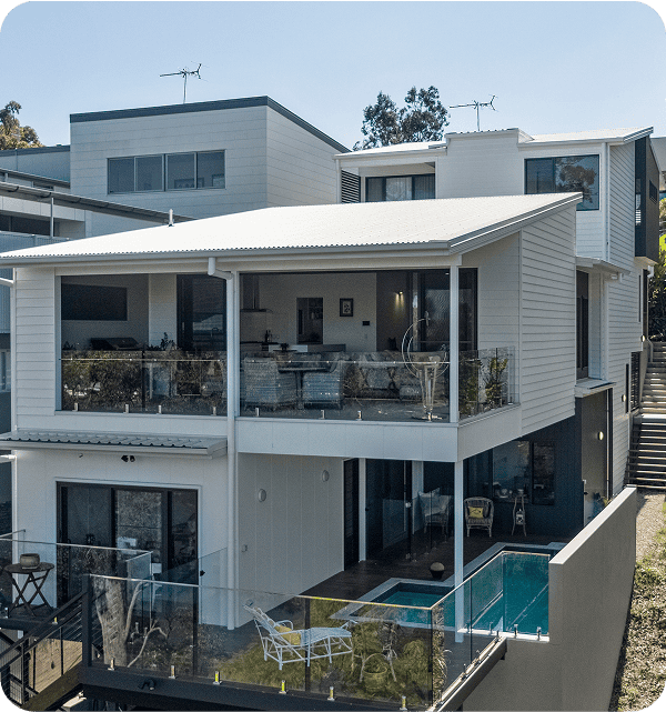 Modern multi-level house with large glass balcony, swimming pool, and outdoor furniture, showcasing contemporary architecture and stylish outdoor living space in a sunny Queensland setting.