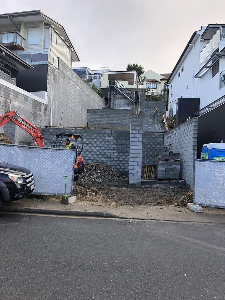 Excavation site for a new residential construction showing retaining walls and construction equipment on a hillside.