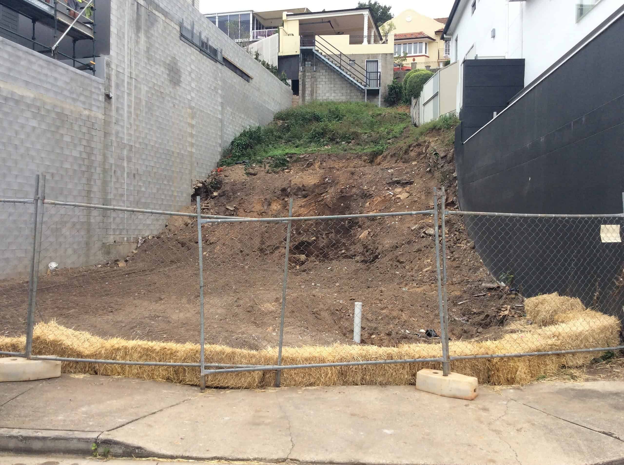 Excavated hillside site with construction fencing, showing an urban slope with mix of exposed soil, concrete retaining walls, and residential homes at the top.