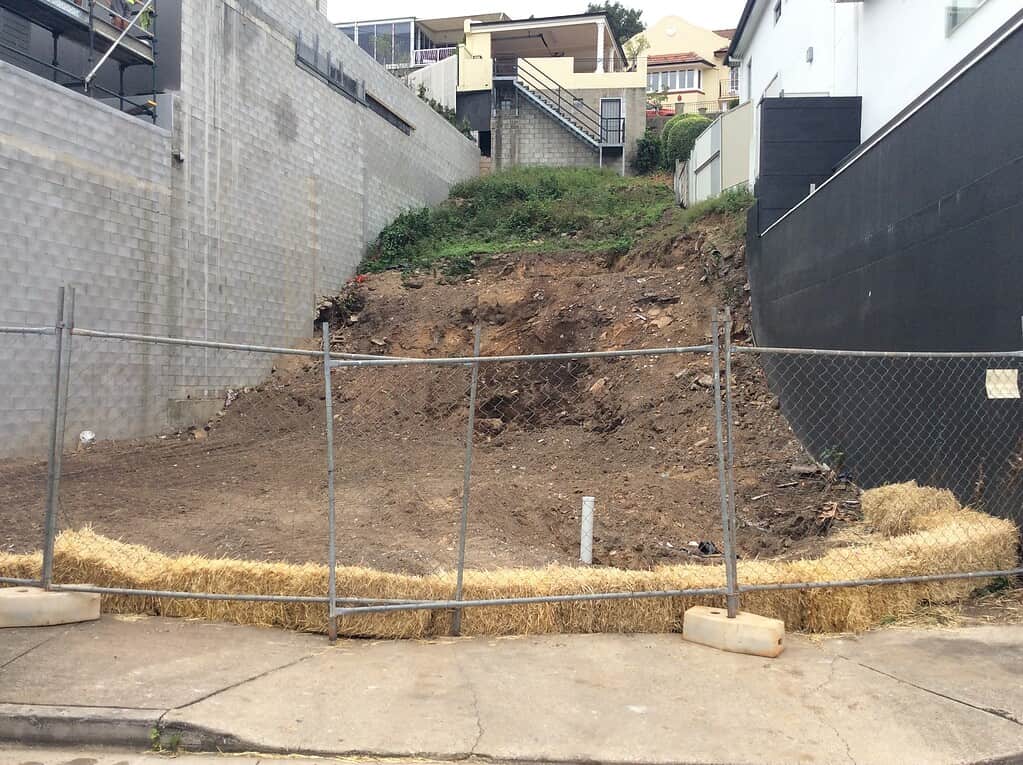 Excavation site for new construction, surrounded by residential houses on an urban slope, with fencing and straw barriers for safety and site management.