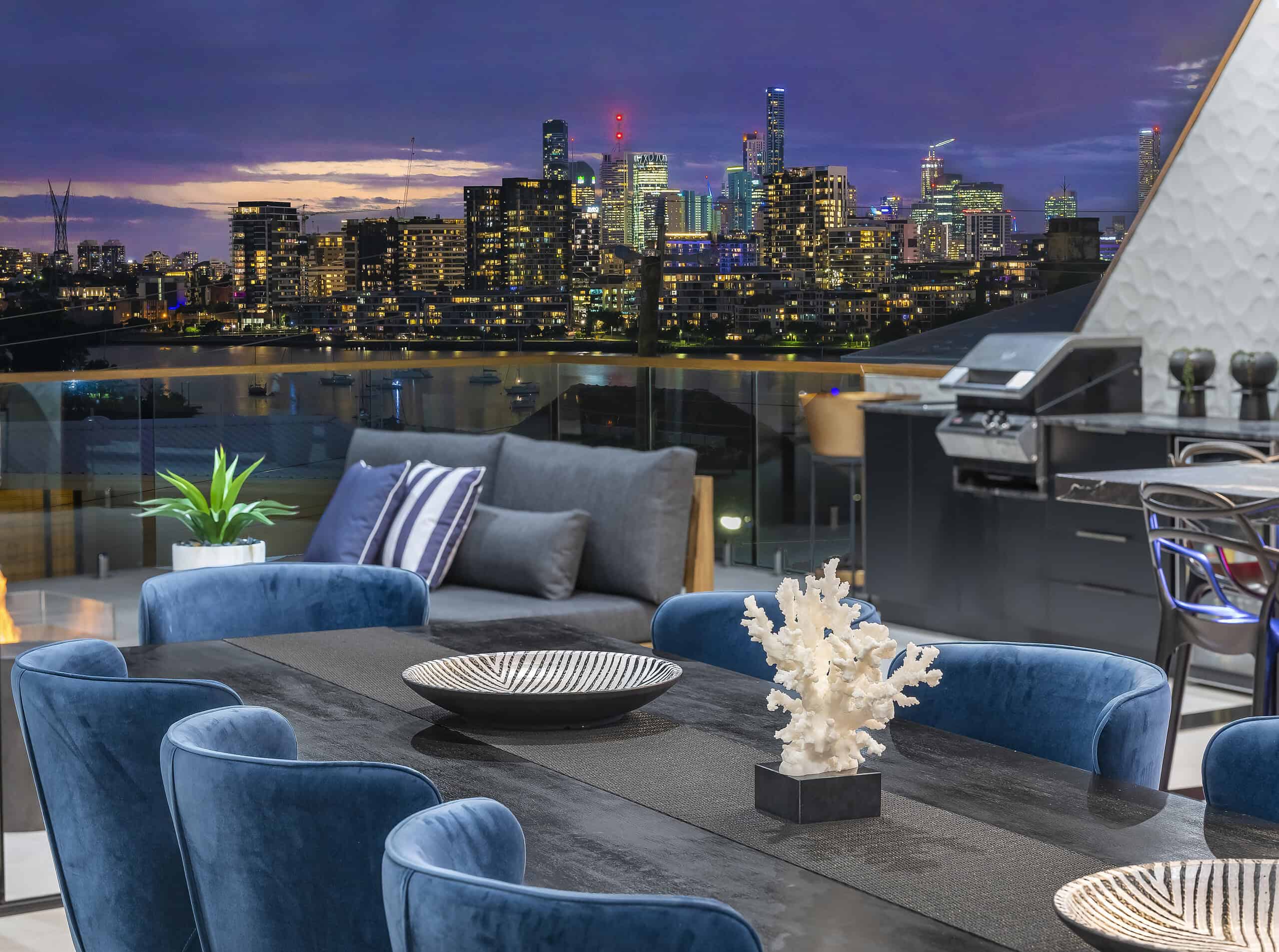Modern rooftop dining area with view of city skyline at dusk, featuring stylish blue chairs, decorative coral centerpiece, and outdoor kitchen setup.