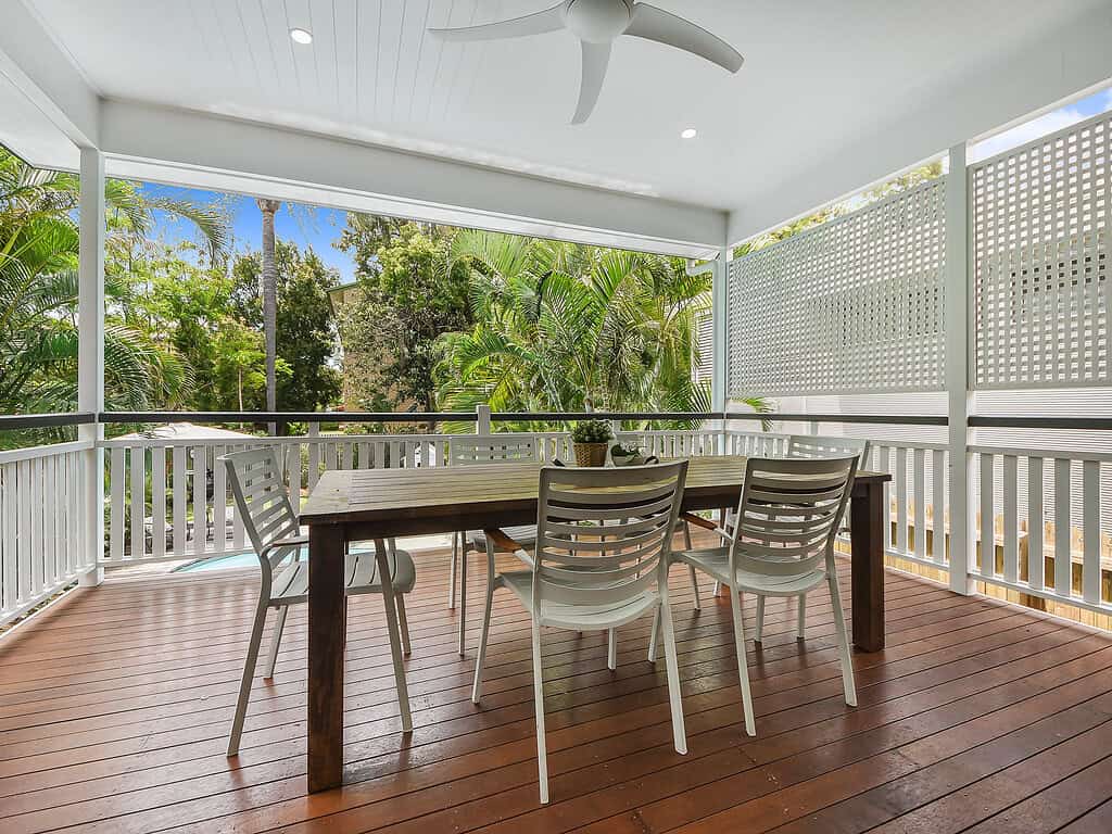 Modern covered outdoor patio with a wooden dining table, white chairs, lush greenery, and a ceiling fan, ideal for relaxing and entertaining in a stylish, tropical backyard setting.