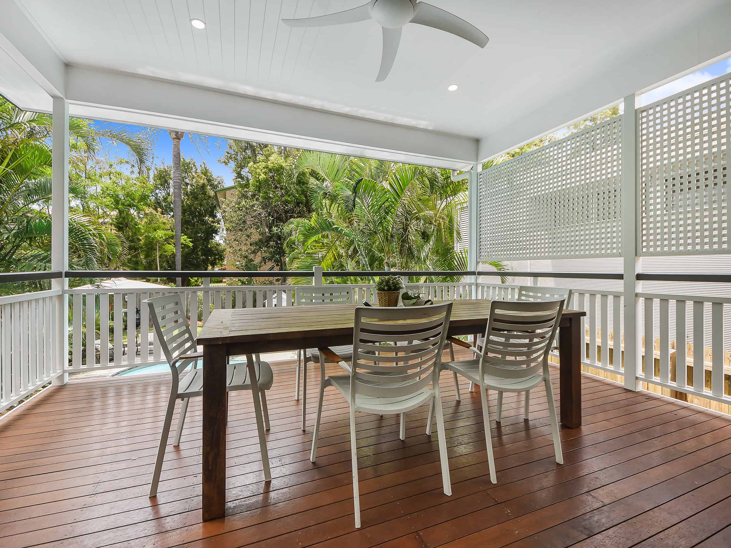Modern covered outdoor patio with a wooden dining table, white chairs, lush greenery, and a ceiling fan, ideal for relaxing and entertaining in a stylish, tropical backyard setting.