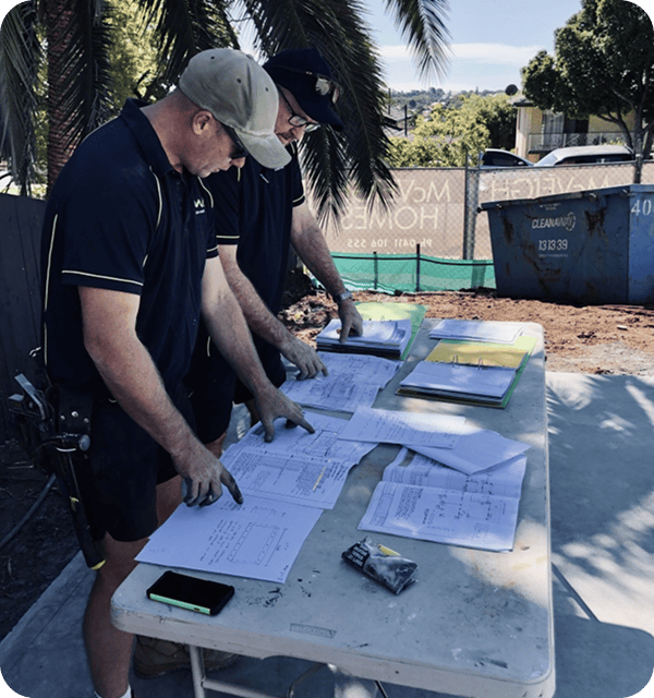 Blueprint review at construction site, workers examining project plans and documents, outdoor setting with palm trees and construction equipment, team collaboration on building project, architectural planning for new structures.