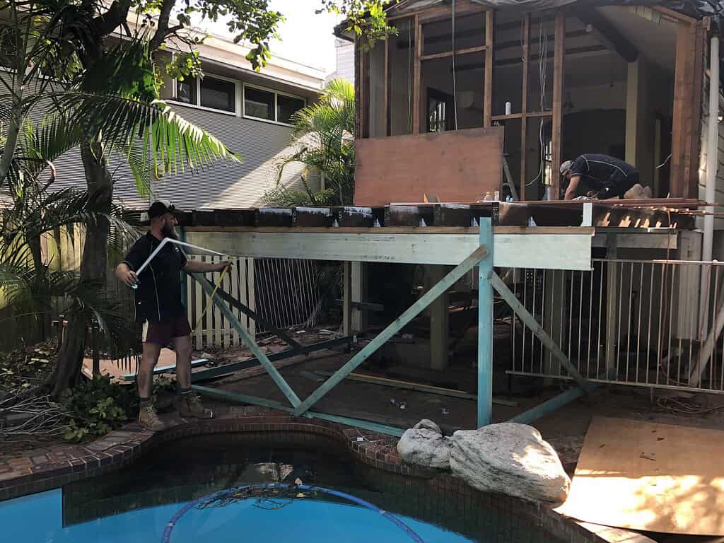 New home construction site with workers building a wooden deck extension over a backyard pool area, showcasing renovation and landscaping progress.