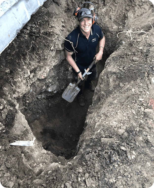 Shovel-wielding worker digging a deep hole outdoors, wearing protective headphones and safety glasses, in a construction or excavation site.