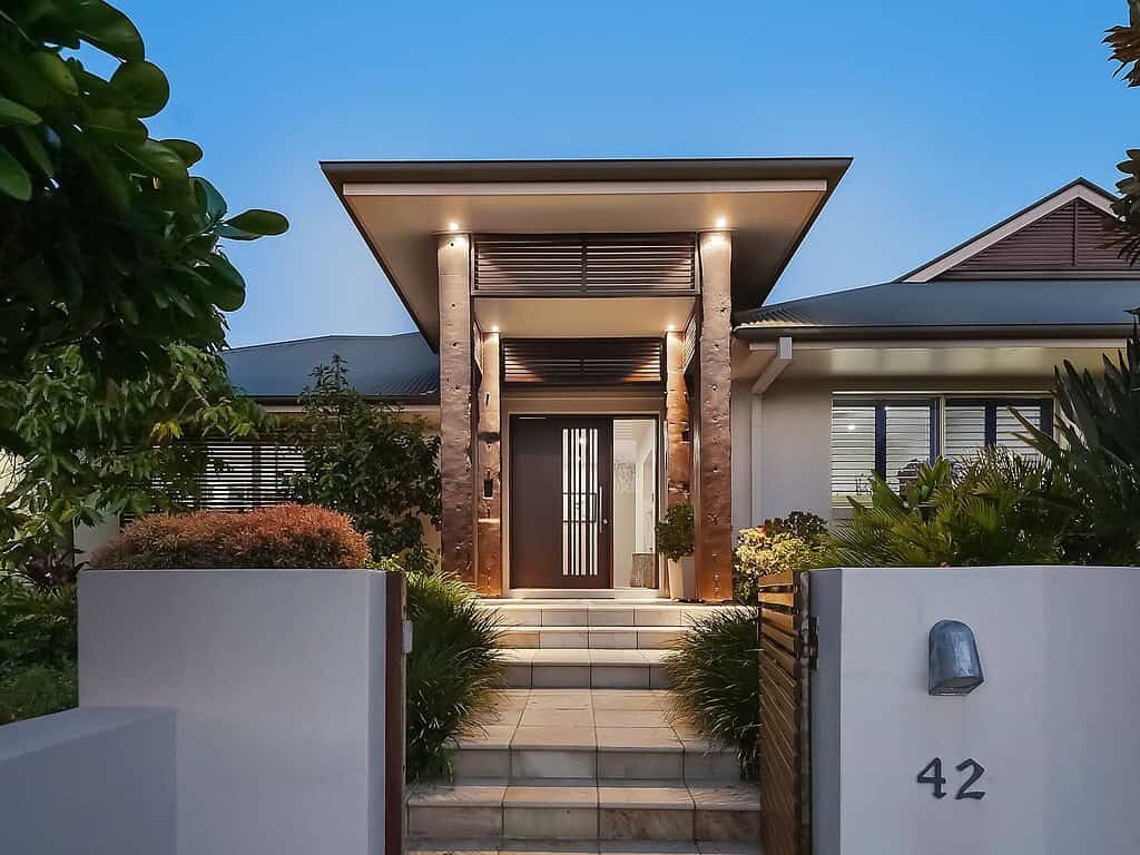 Modern residential house entrance with stylish wooden accents and welcoming lighting, surrounded by lush greenery, showcasing elegant home design and landscaping.