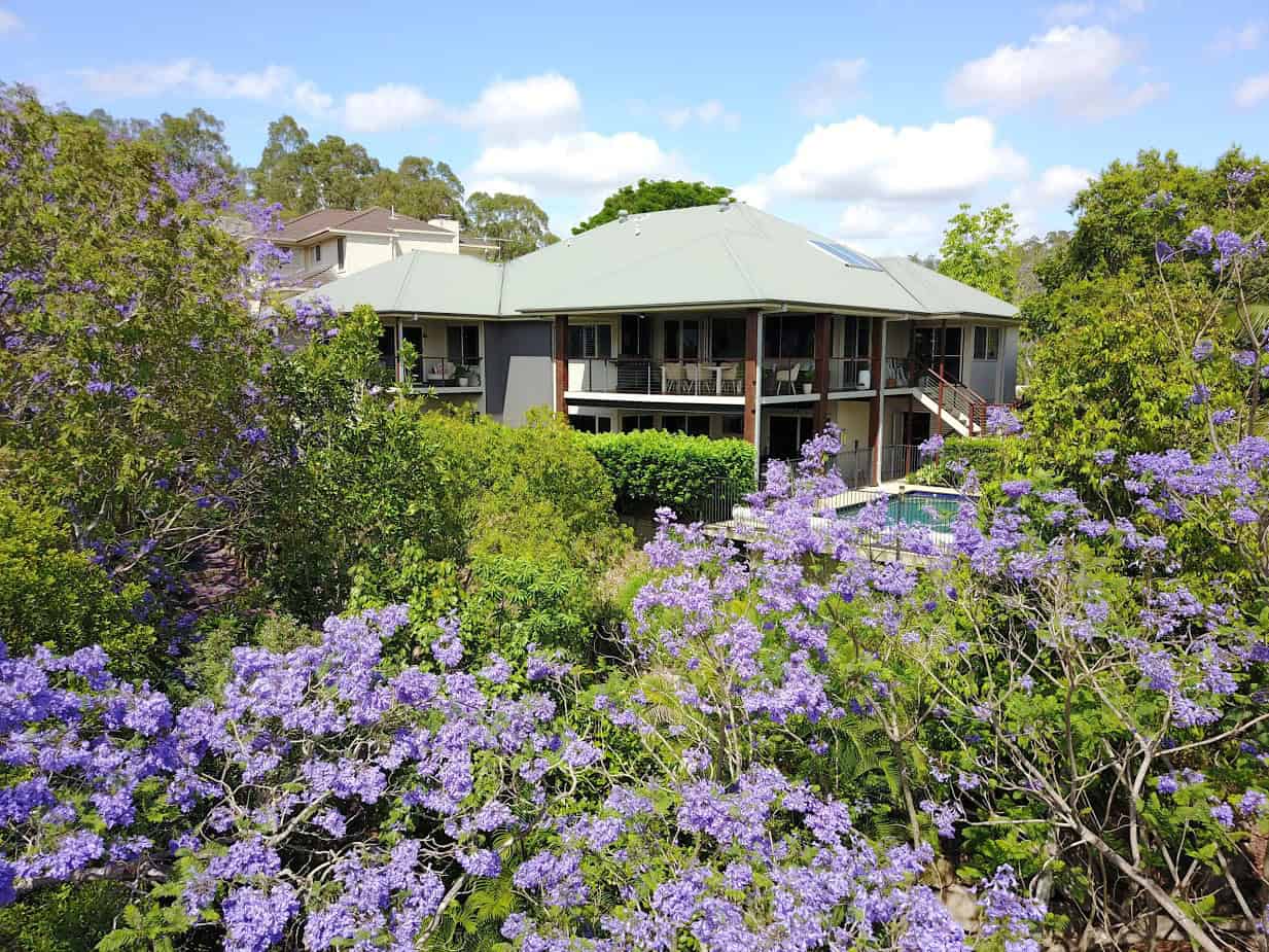 Modern house with a balcony surrounded by lush green trees and vibrant purple flowers on a sunny day, showcasing outdoor living and landscaping.