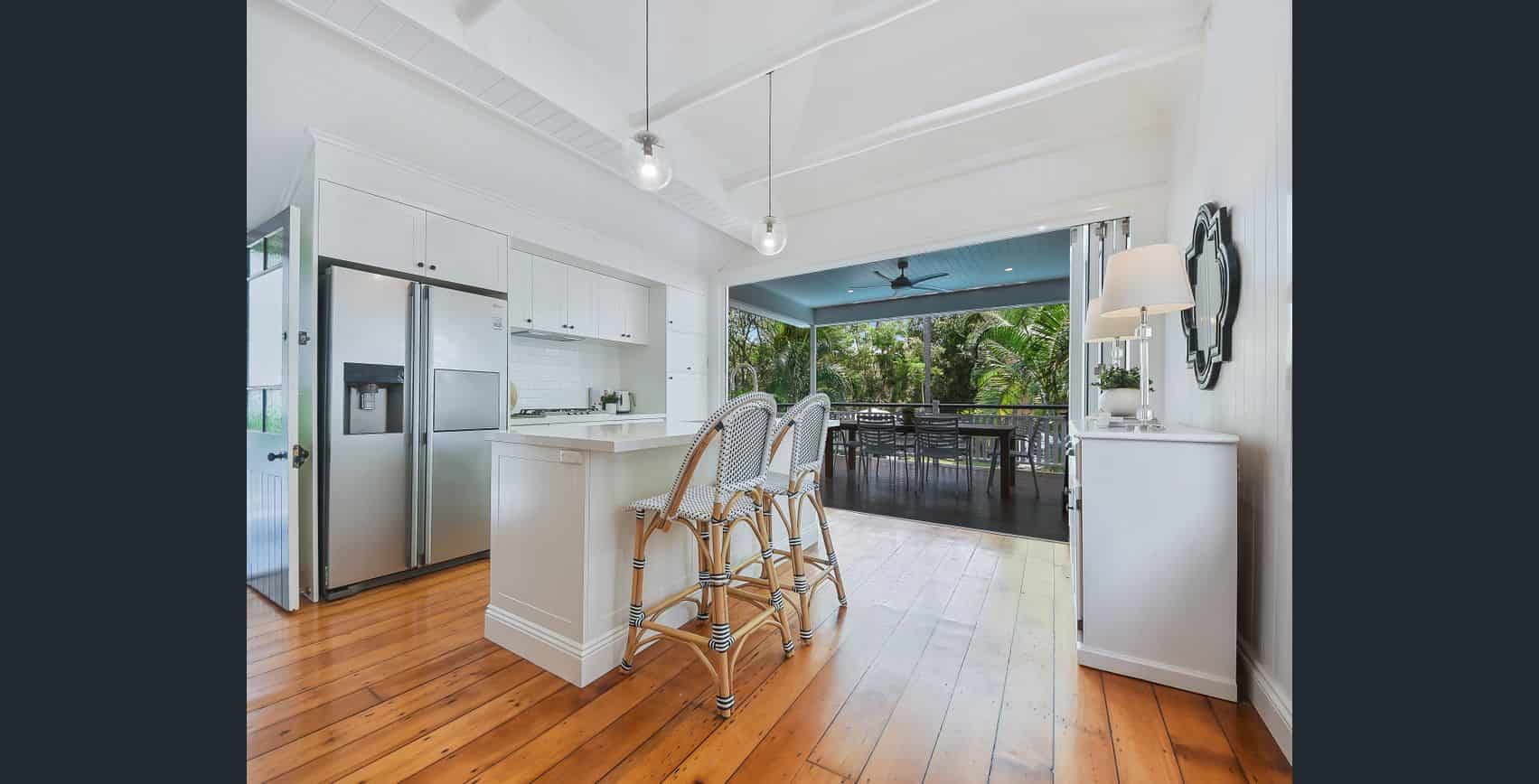 Stunning modern kitchen and outdoor dining area in a newly built home, featuring white cabinetry, stainless steel appliances, wooden flooring, and a spacious screened-in veranda with lush greenery.